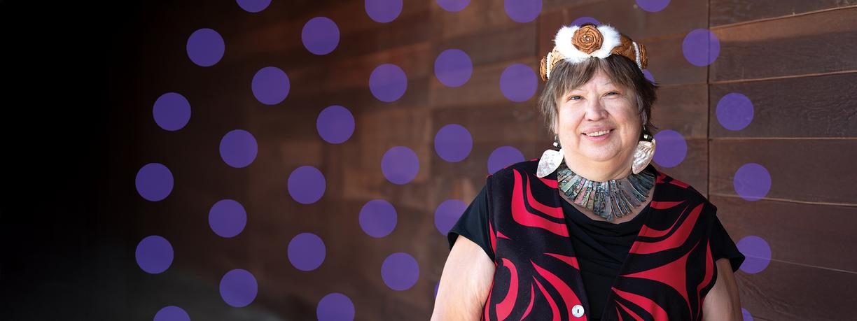 Smiling woman wearing traditional Indigenous attire in front of purple dots