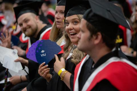 a graduate holds a fan that says VIU and points to it while graduates on either side of her look on