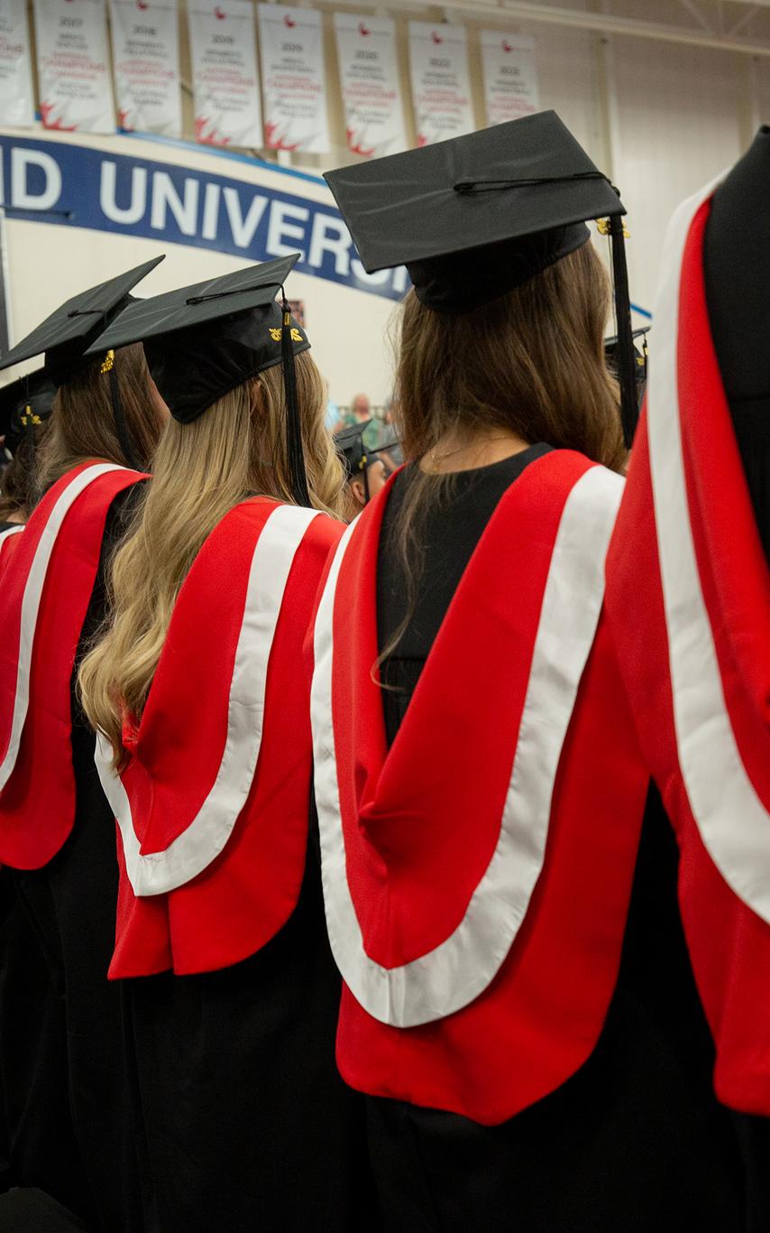 graduates in their regalia with red stoles sit in chairs at convocation